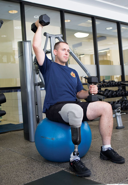 An Afghanistan war amputee veteran sits on an exercise ball while lifting weights at a gym.