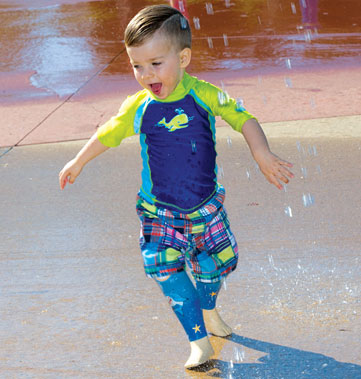 A young male double leg amputee plays in the water at a splash pad.