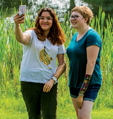 Two teenage females, one an arm amputee and one a hand amputee, stand in front of a pond and take a selfie.