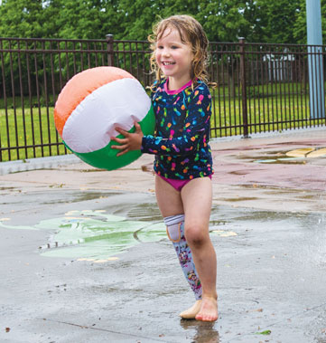A young female with a Syme’s amputation holds a beach ball while playing in the water at a splash pad.