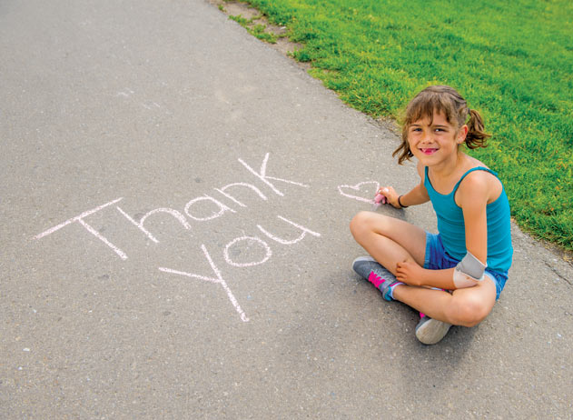 A young amputee girl wearing a prosthetic arm writing “thank you” in chalk on a sidewalk.