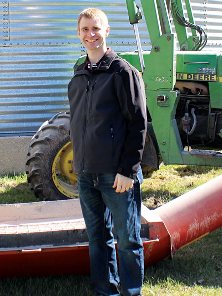 An adult male with a shoulder disarticulation stands in front of a grain auger and a tractor. 