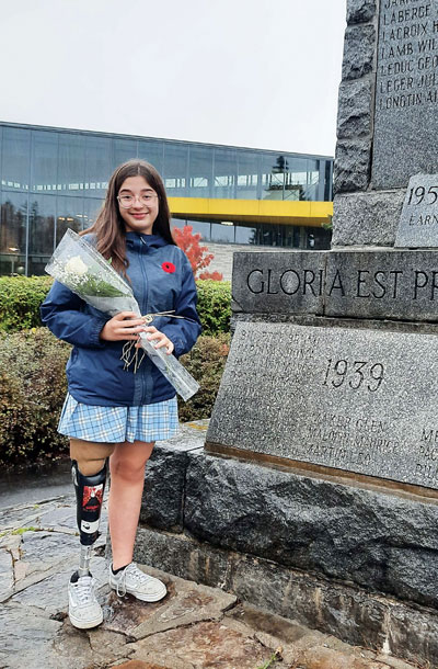 A young female leg amputee stands beside a cenotaph and is holding a white rose.