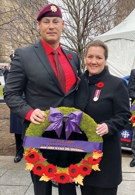 An adult male double leg amputee holds a War Amps Remembrance Day wreath with his wife.