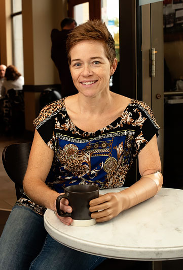A female adult arm amputee sits at a cafe table and holds a mug.