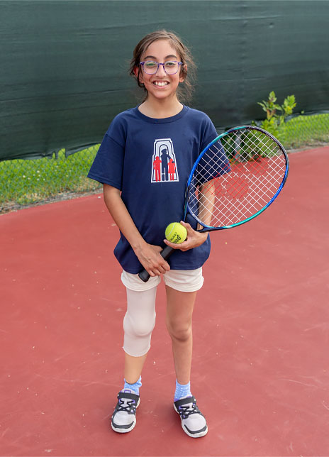 Une jeune fille amputée d’une jambe tient une raquette et une balle de tennis en étant debout sur un terrain de tennis.