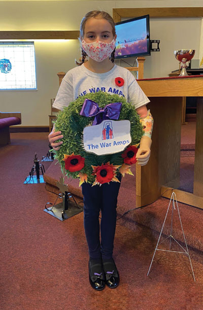 A young female arm amputee holds a Remembrance Day wreath with The War Amps written on it while wearing a cloth face mask.
