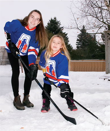 Audrey and Leah standing by an outdoor rink demonstrating how they hold their hockey sticks with their specialized artificial arms.