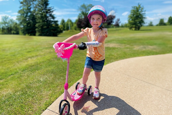 A young female arm amputee smiles while riding a scooter using a device that attaches to the handlebar.