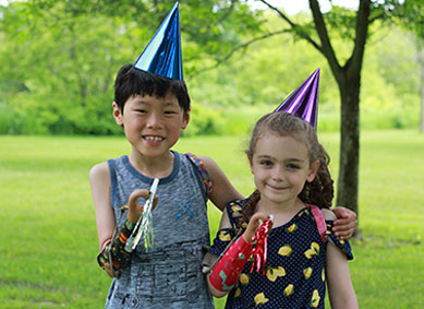 Two young arm amputees standing in a park with their arms around each other wearing party hats and holding party horns.